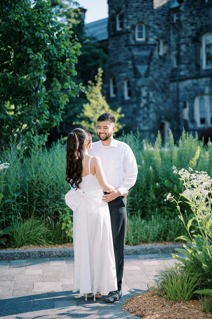 A couple in formal attire smiles and embraces in a picturesque garden setting, with a historic stone building in the background. The woman wears a white dress with a large bow, and the man is in a white shirt and dark pants. Lush greenery and flowering plants surround them under bright daylight.
