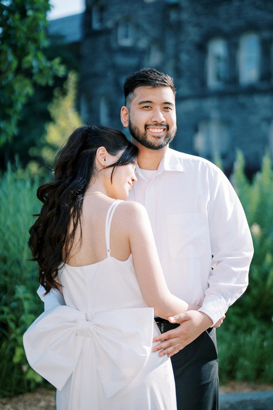 A happy couple embraces outdoors, dressed in white attire, with a lush green garden and stone building in the background, capturing a romantic moment.