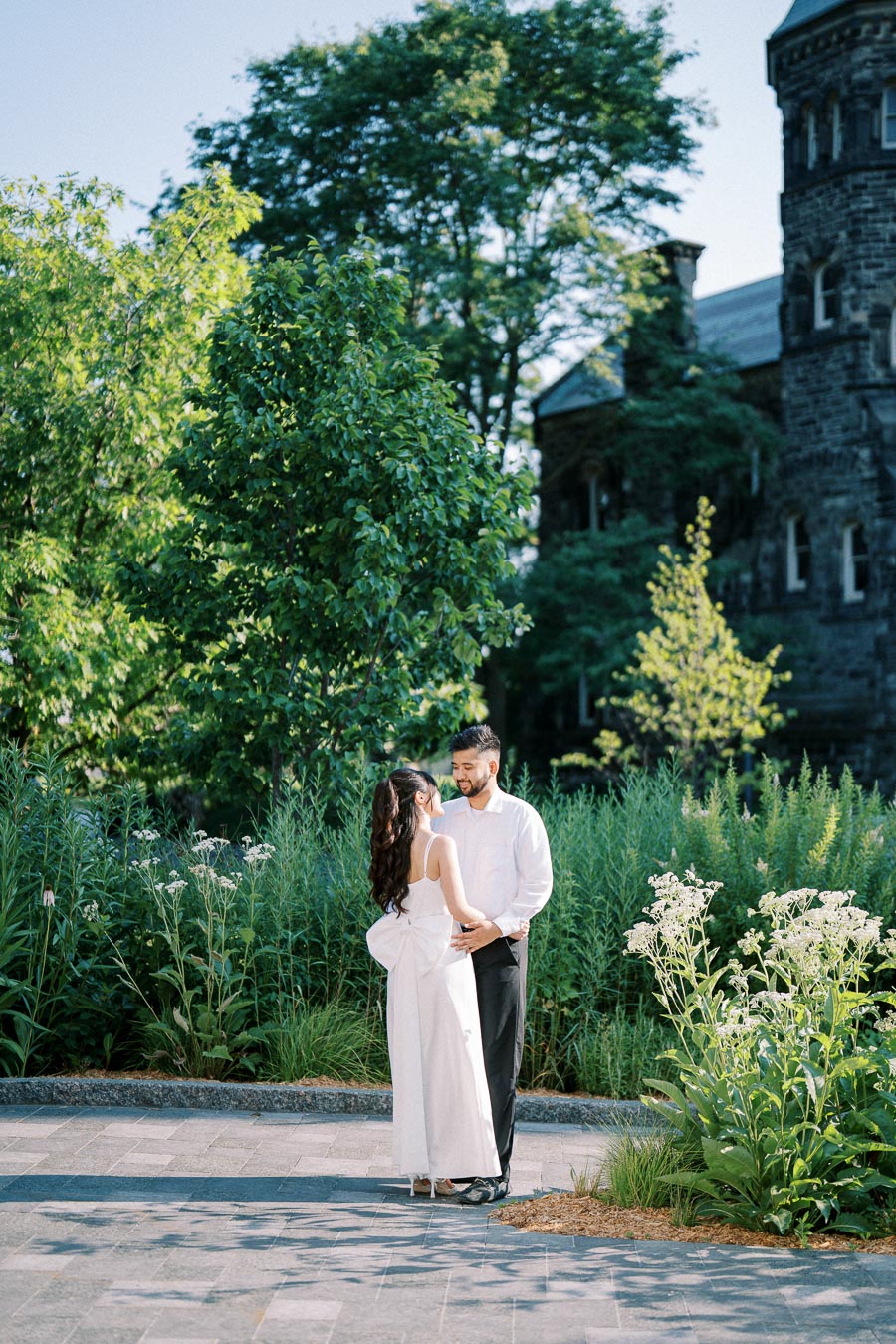 A couple embracing in a lush garden setting with greenery and historic stone building in the background, captured on a sunny day.