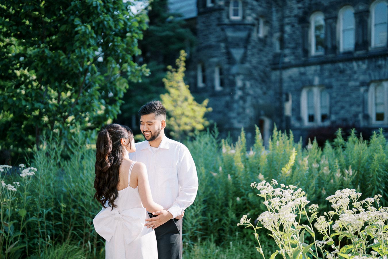 A joyful couple smiling at each other in a garden setting, surrounded by lush greenery and flowers, with a historic stone building in the background.