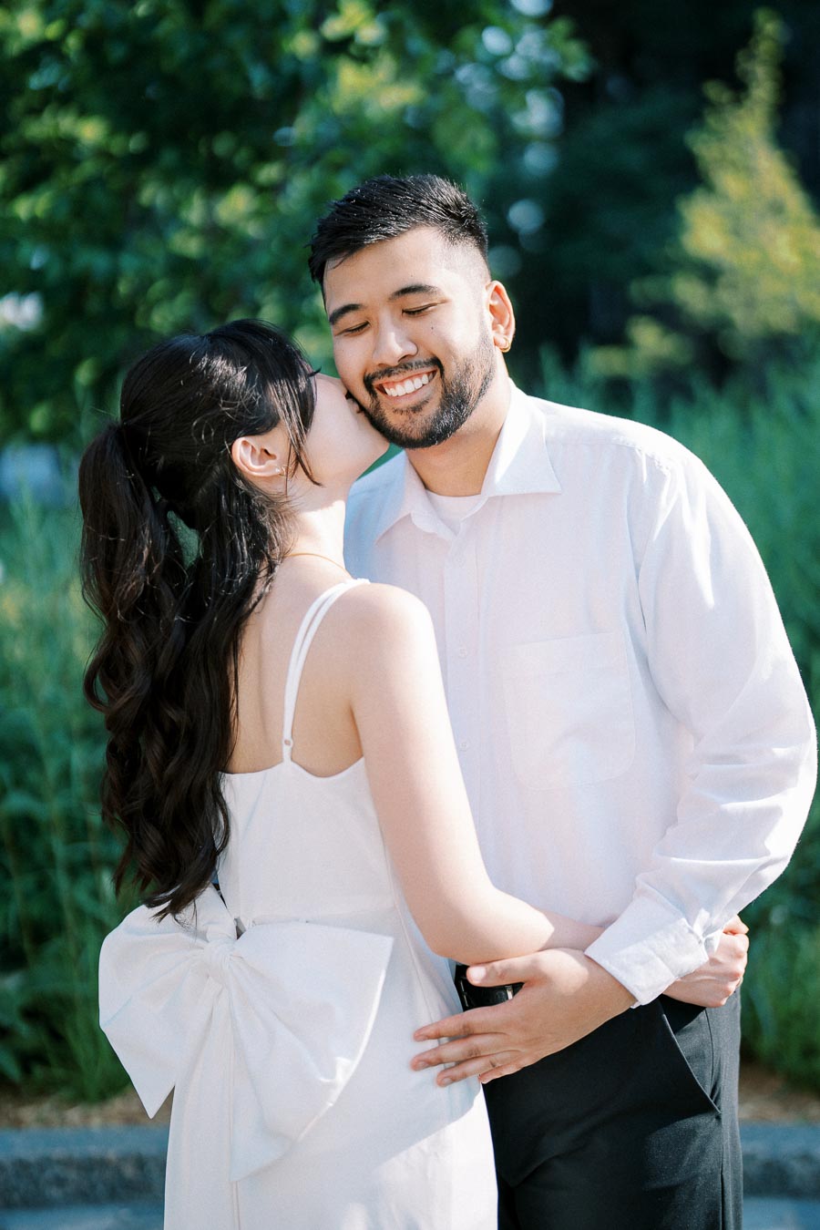 A couple embracing outdoors, with a woman in a white dress and bow kissing a man in a white shirt, both smiling, in a green, sunny garden setting.