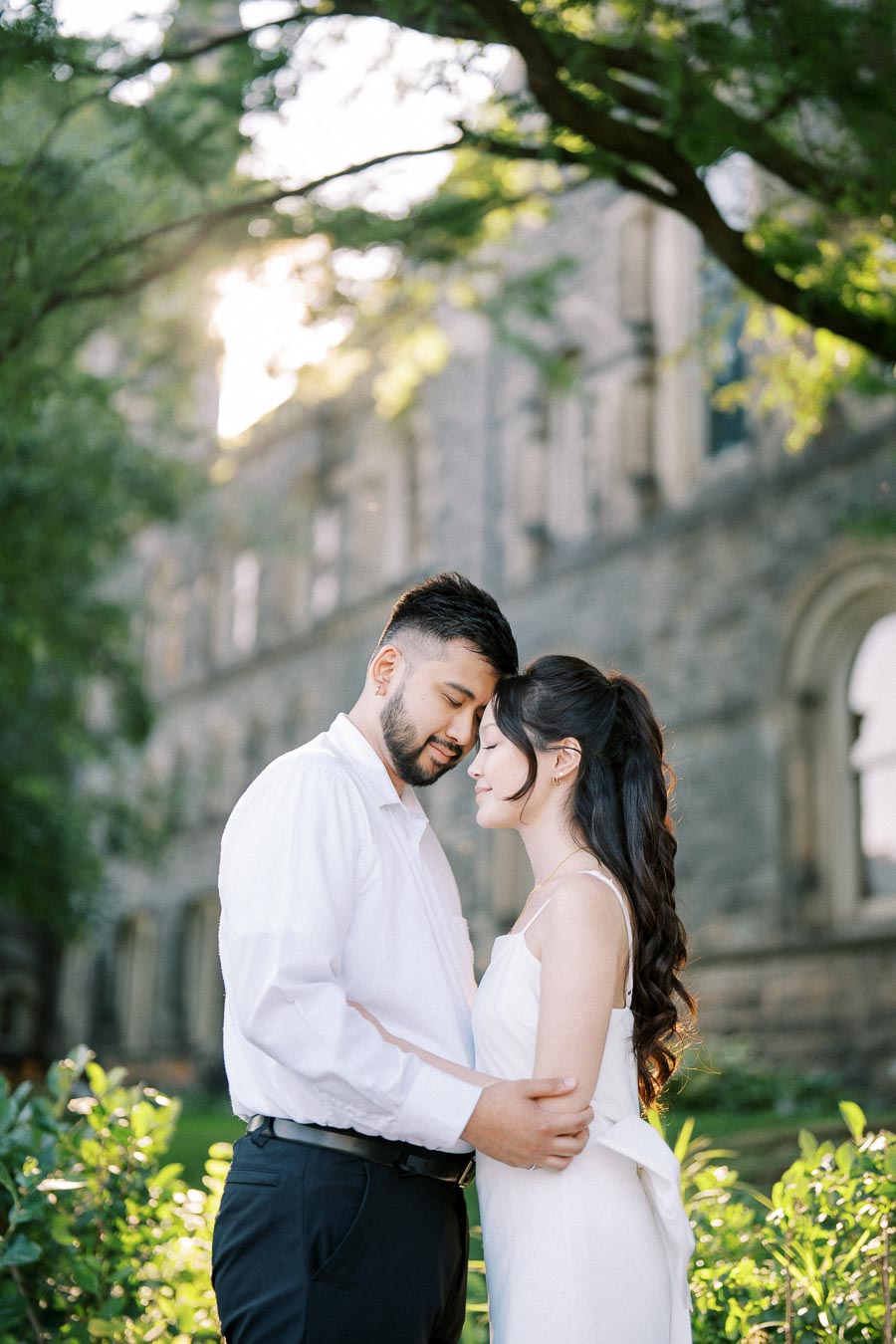 A couple in white attire embraces warmly in a sunlit garden, with a historic stone building in the background.
