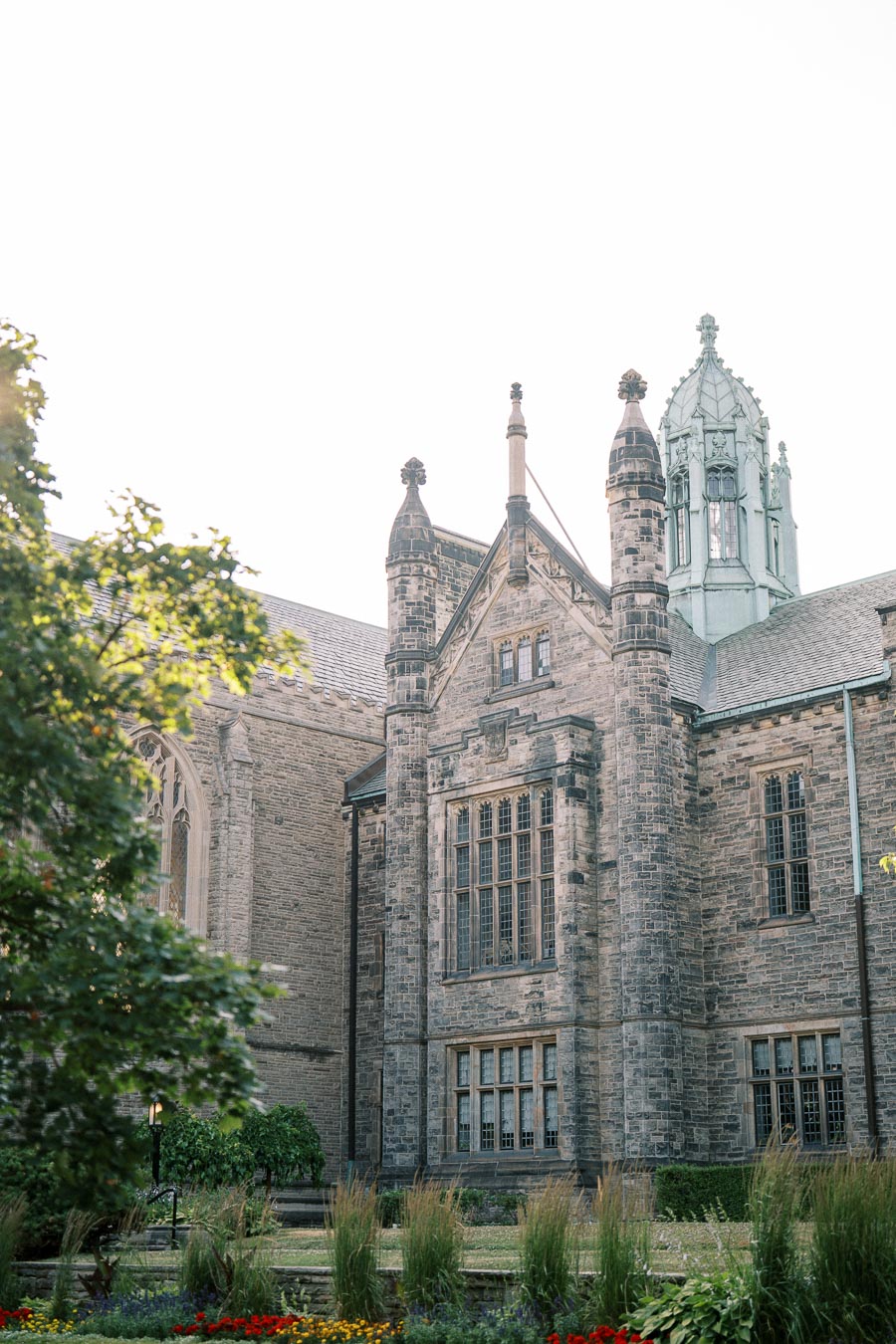 Historic stone building with ornate architecture and tower against a bright sky, surrounded by lush greenery and colorful flower beds in the foreground.