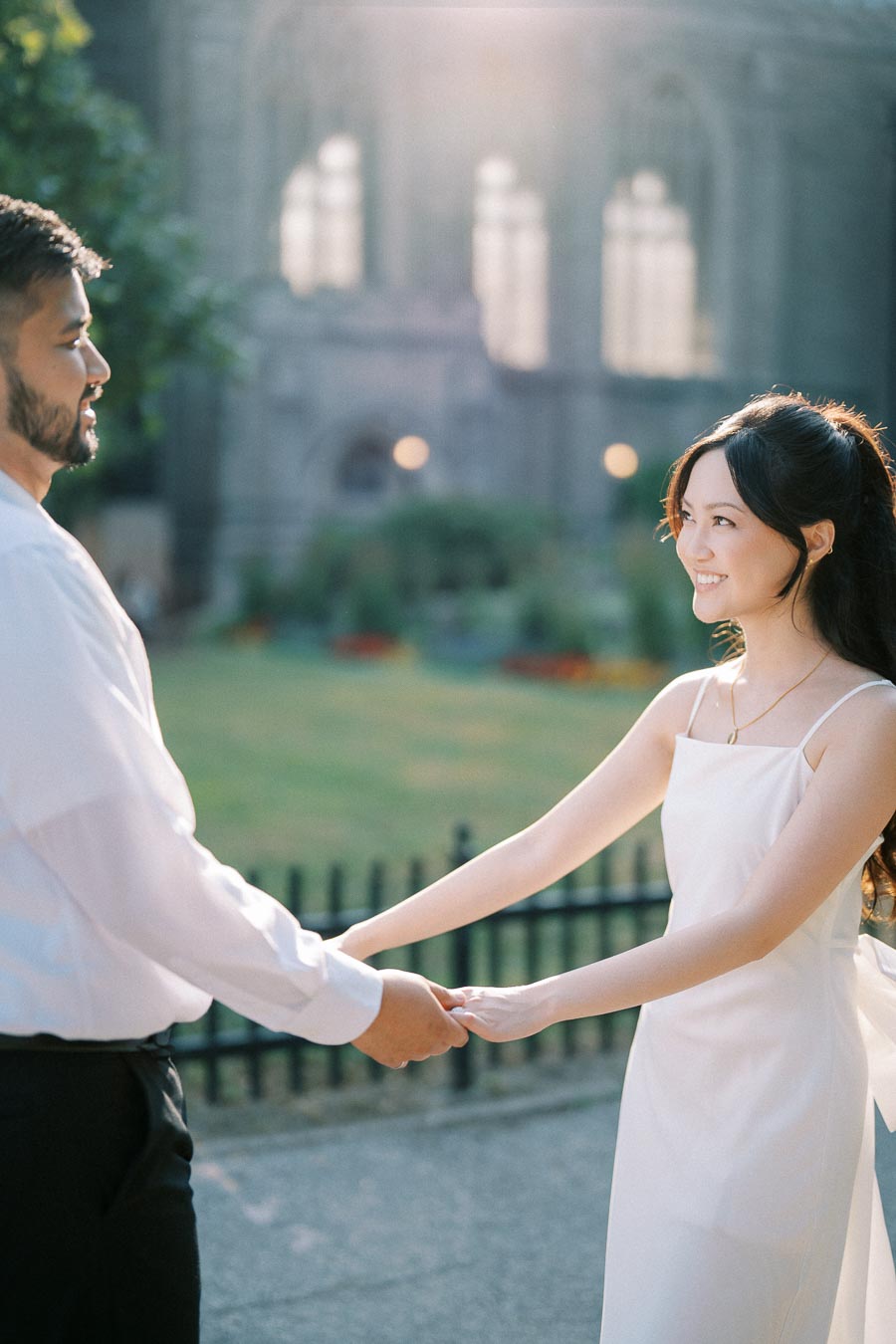 A couple holding hands outdoors, surrounded by greenery and soft sunlight. The woman is wearing a white dress, and the man is dressed in a white shirt, creating a serene and romantic atmosphere. Ideal for engagement or wedding-themed content.