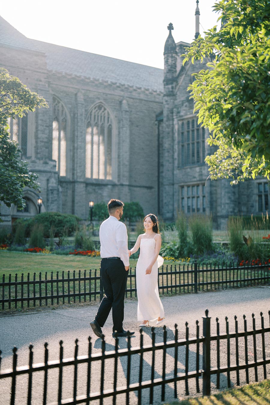 A couple in formal attire holds hands near a historic stone building, surrounded by lush greenery and a black wrought iron fence. The sun casts a soft glow, creating a romantic ambiance.