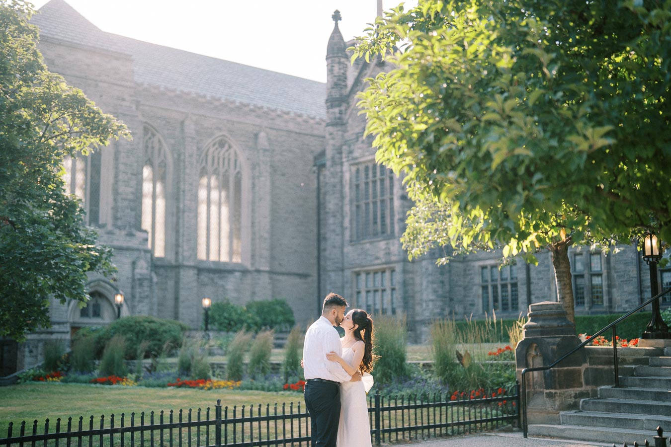A romantic couple embracing in a garden setting with a historic stone building in the background, surrounded by lush greenery and vibrant flowers.