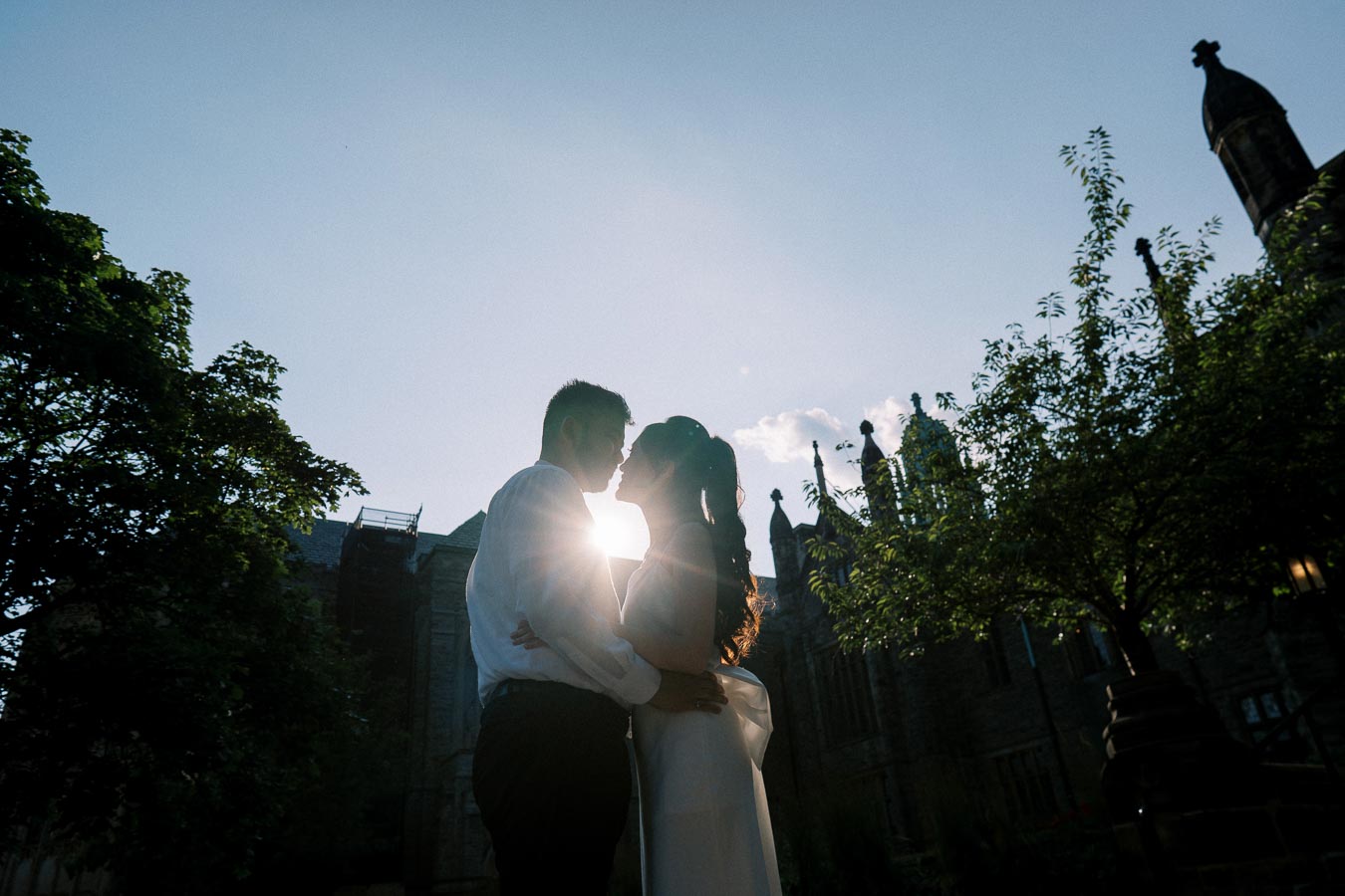 Silhouette of a couple embracing at sunset in front of a historic stone building, surrounded by trees, with lens flare highlighting the romantic atmosphere.