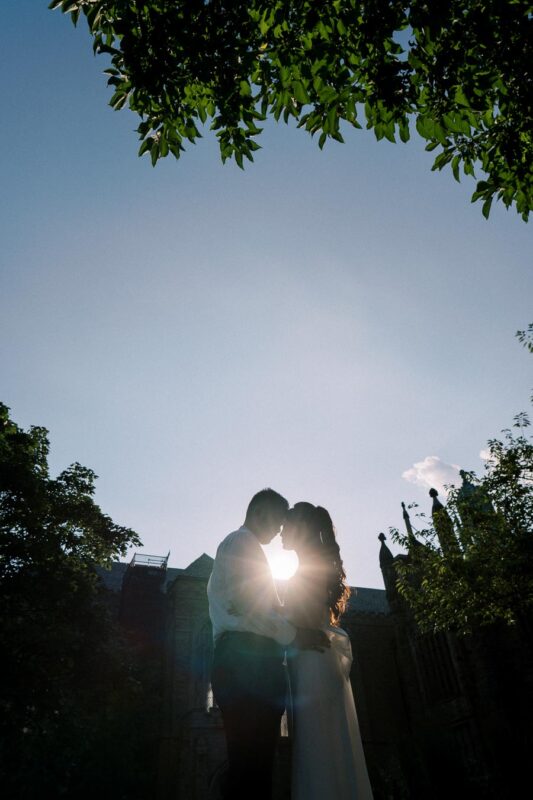 Silhouette of a couple embracing under a tree, with the sun creating a romantic glow behind them, set against a clear blue sky.