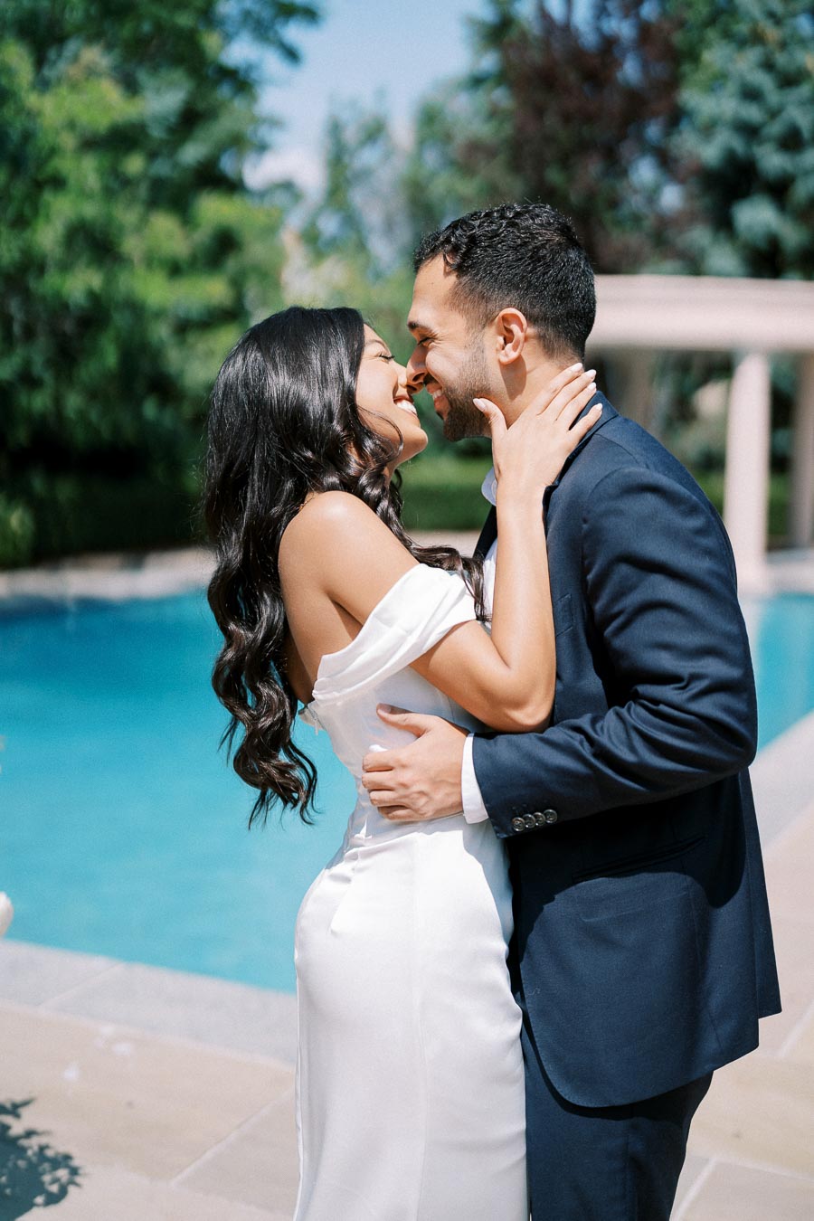 Romantic couple embracing near a swimming pool with lush greenery in the background, both dressed elegantly in formal attire for a special occasion.