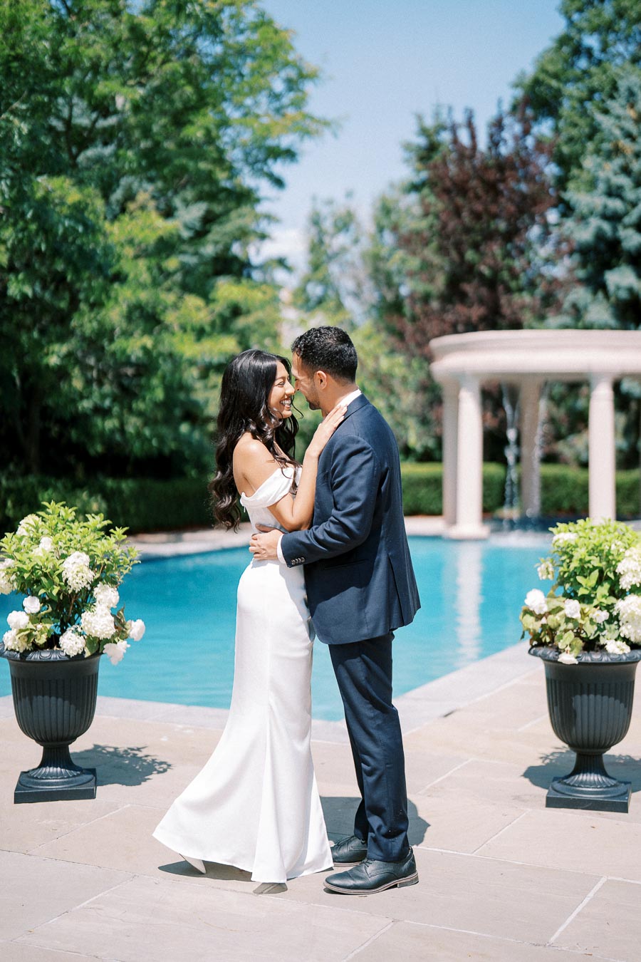 A couple embracing by a poolside, surrounded by lush greenery and elegant floral arrangements, under a clear blue sky.