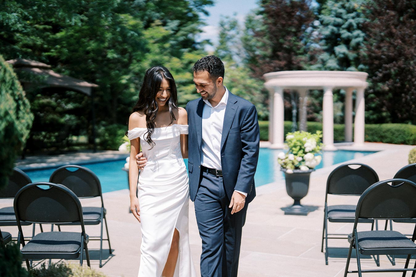 Happy couple walking arm in arm by a poolside venue, dressed in elegant wedding attire, surrounded by lush greenery and outdoor seating.