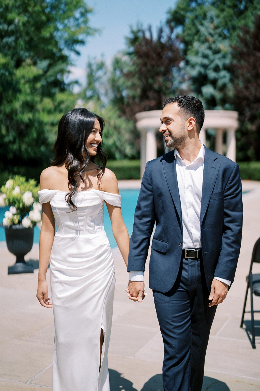 Couple Holding Hands and Smiling in Sunlit Outdoor Setting, Woman in Elegant White Dress and Man in Dark Suit