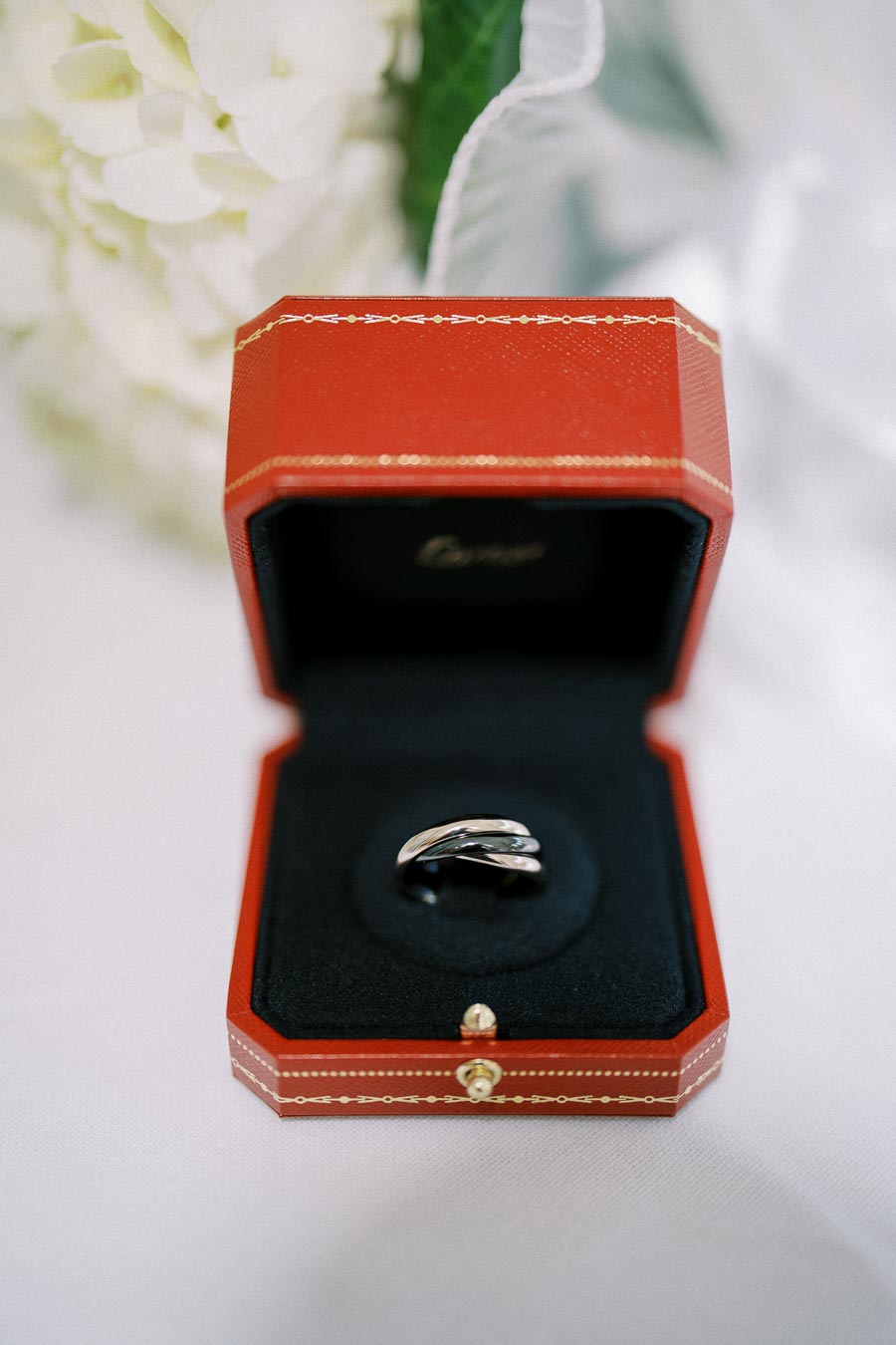 Elegant silver ring in a red jewelry box with a black velvet interior, placed on a white surface, surrounded by soft-focus white flowers in the background.