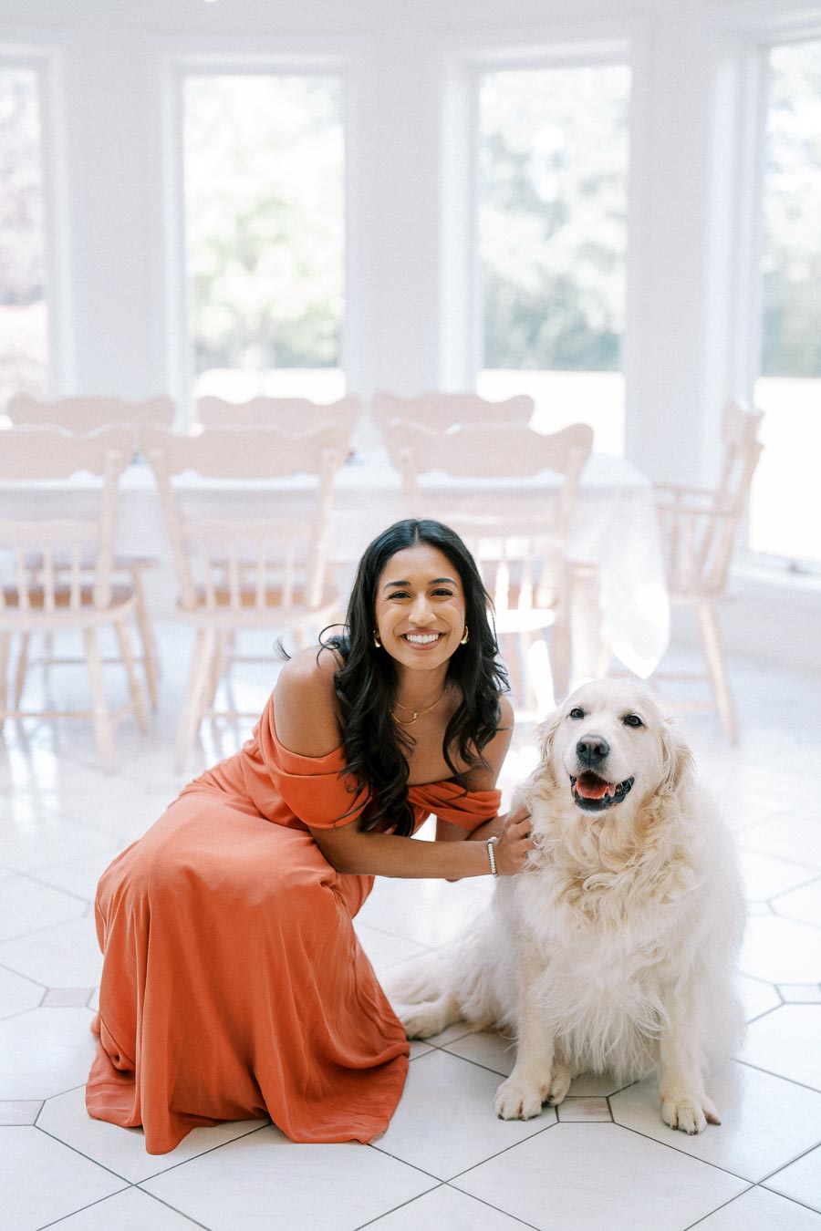 A woman in an orange dress smiling and kneeling beside a fluffy white dog in a bright room with large windows and wooden chairs.