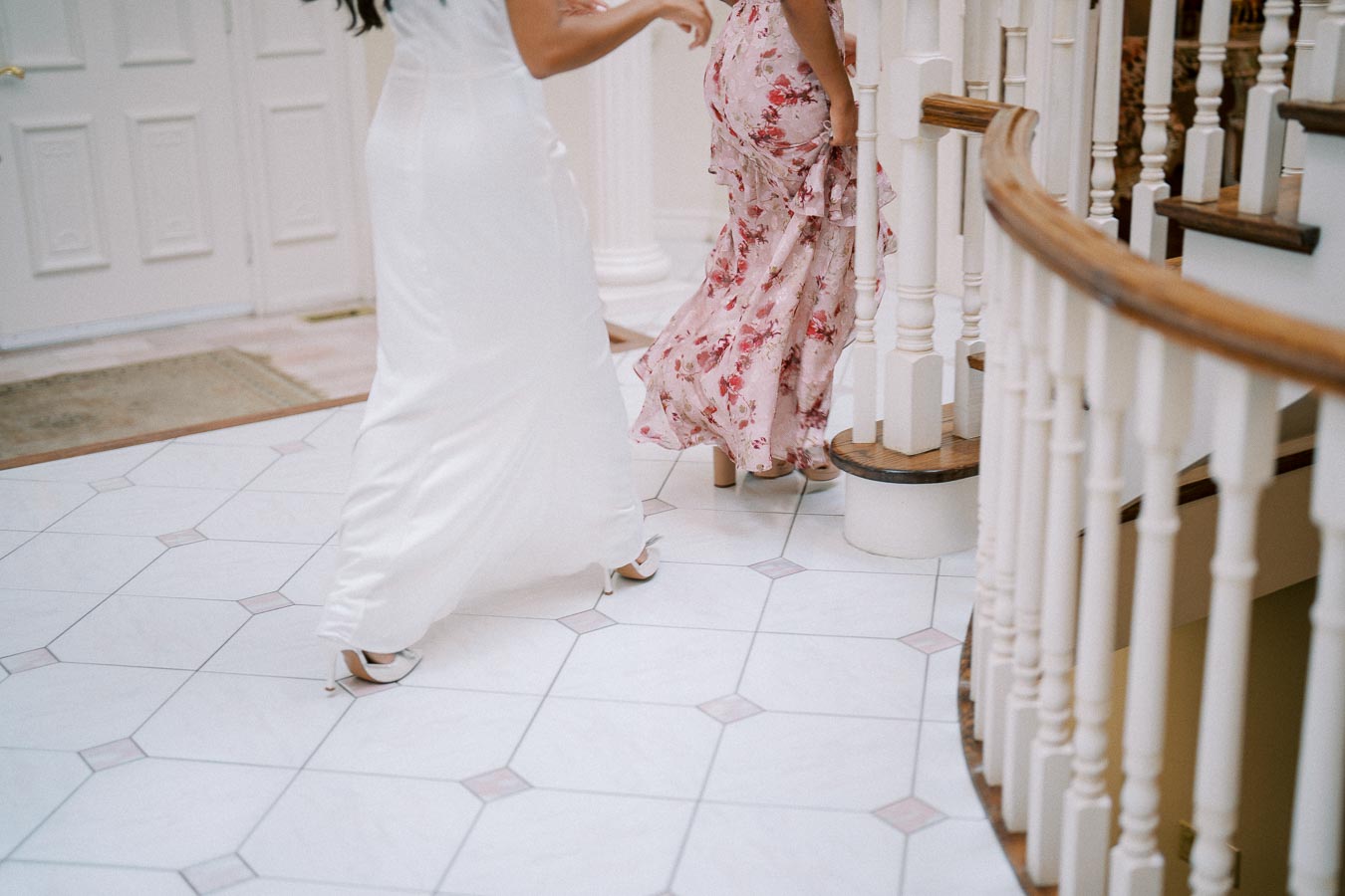 Two women in elegant dresses ascending a spiral staircase, one in a white gown and the other in a floral pink dress, showcasing wedding or formal event fashion.