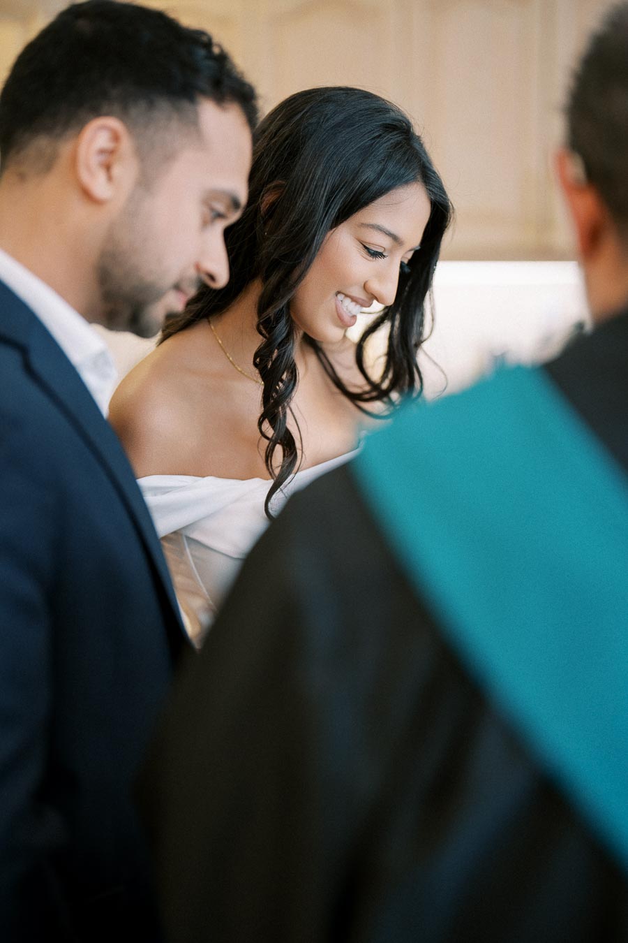 A woman smiling warmly during a formal indoor gathering, accompanied by two other people, all dressed in elegant attire.