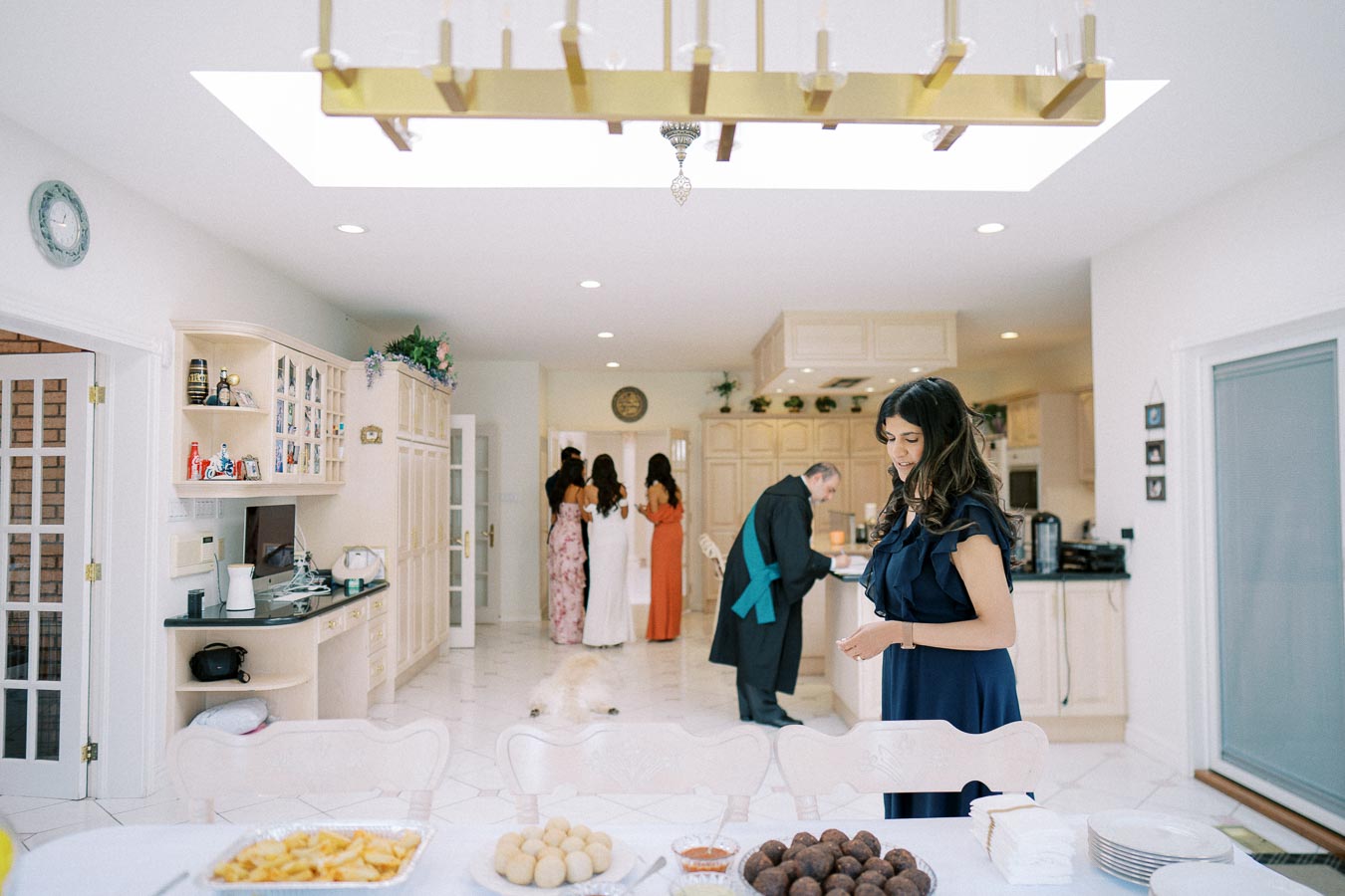 Elegant kitchen setting with a modern chandelier and bright lighting, featuring a woman in a navy dress preparing for a gathering; in the background, a man wearing a robe and other women in colorful dresses. A variety of snacks, including fried treats and sauces, are placed on the table, ready for guests.
