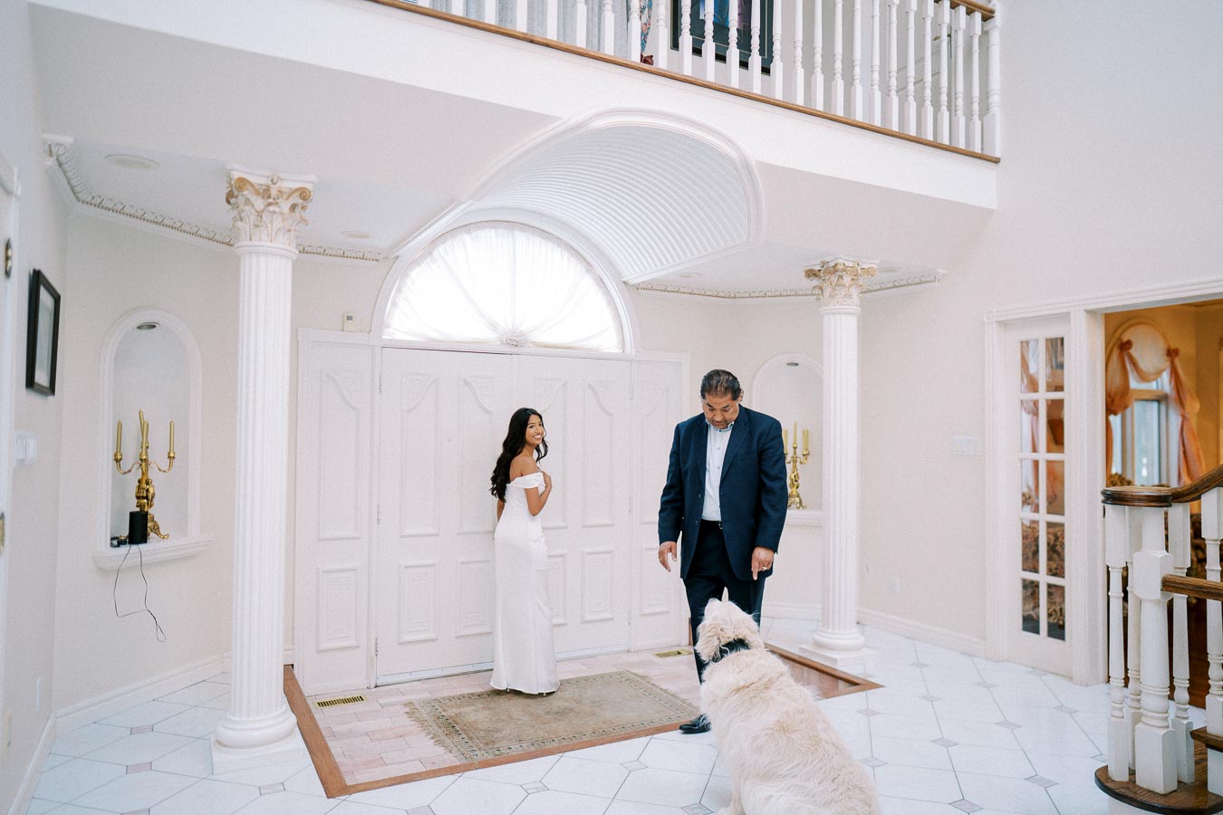 Elegant indoor setting with a woman in a white dress and a man in a suit interacting with a large white dog, surrounded by classic white columns and luxurious decor.