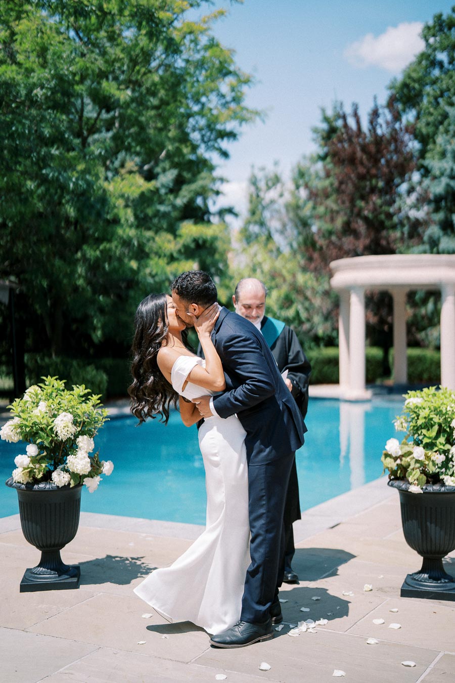 A couple shares a passionate kiss at their outdoor wedding ceremony by a pool, surrounded by lush greenery and elegant floral arrangements. The bride wears an elegant white gown, and the groom is in a classic navy suit. A serene and romantic setting captures the joy of their special day.