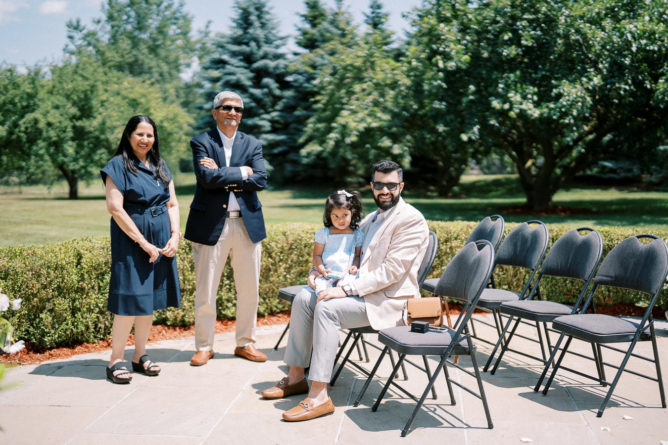 A family enjoying a sunny day outdoors in a garden setting, with two adults standing and a father sitting with his young daughter on a row of chairs.