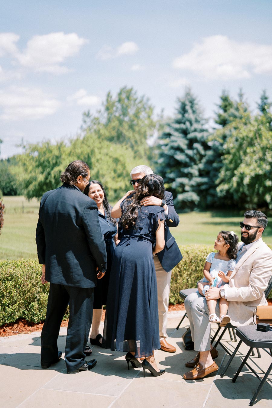 A group of people socializing outdoors at a formal event, with greenery and trees in the background.