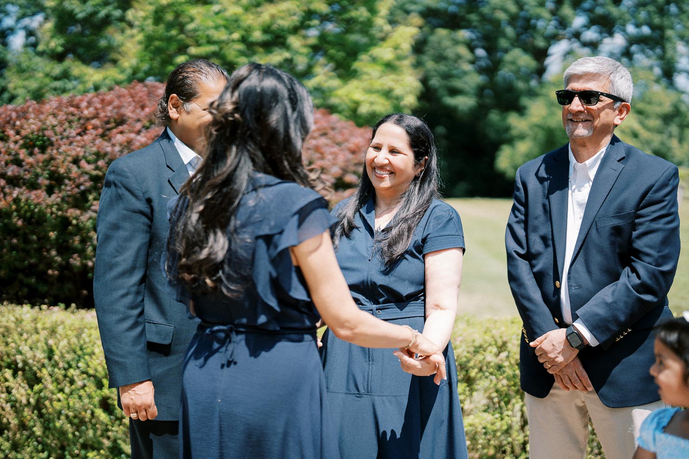 A group of people happily interacting outdoors, dressed in formal attire on a sunny day, standing in a garden setting with green foliage and manicured bushes in the background.