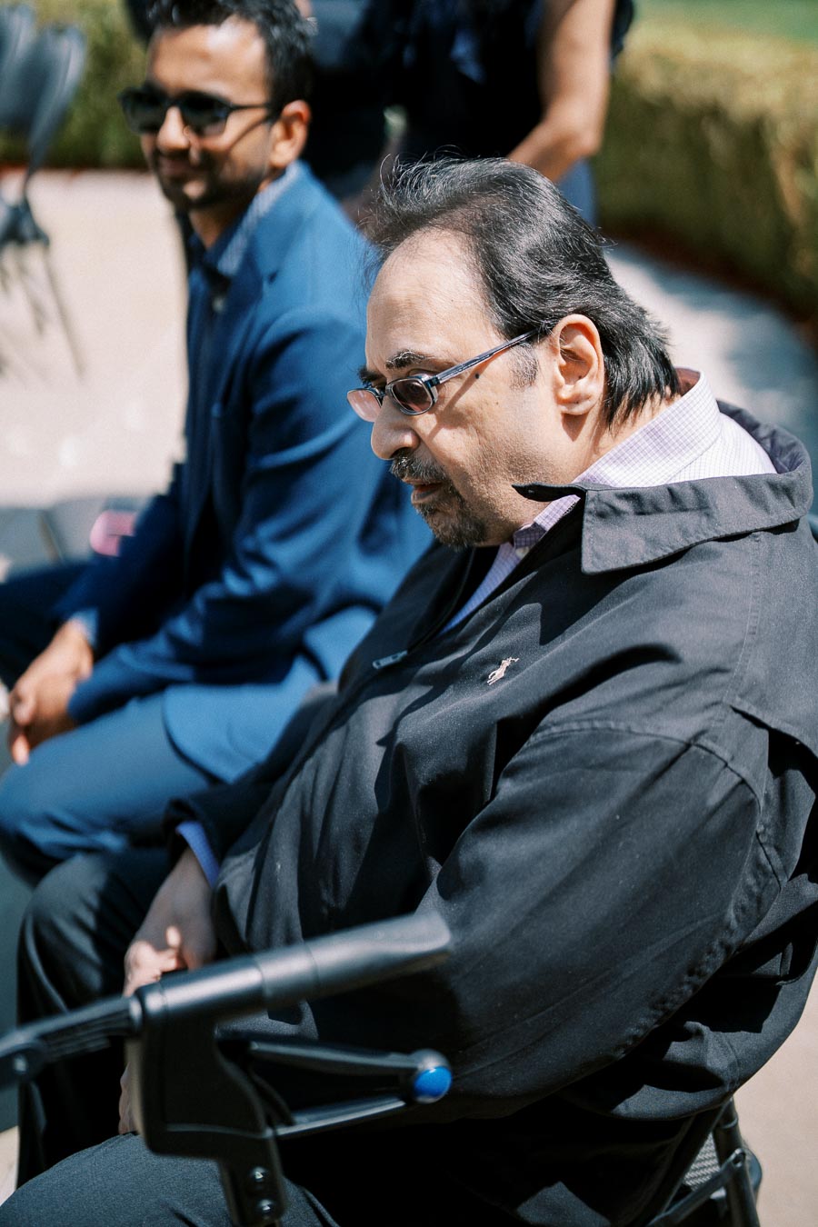 A man outdoors sitting beside another person, using a mobility aid, wearing glasses, and dressed in a black jacket.