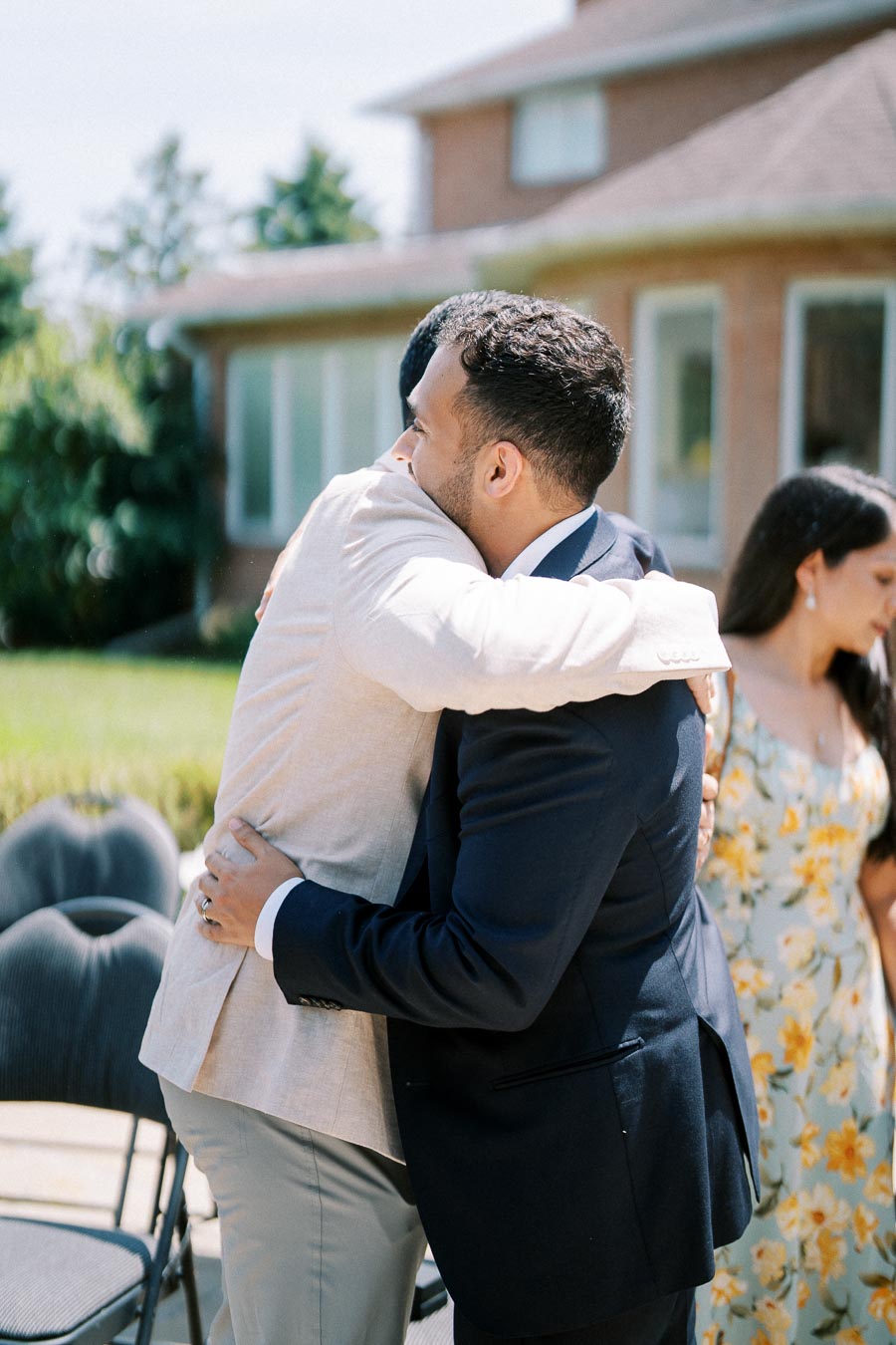 Two men embracing in a friendly hug outdoors at a social gathering, with a woman in a floral dress standing nearby.