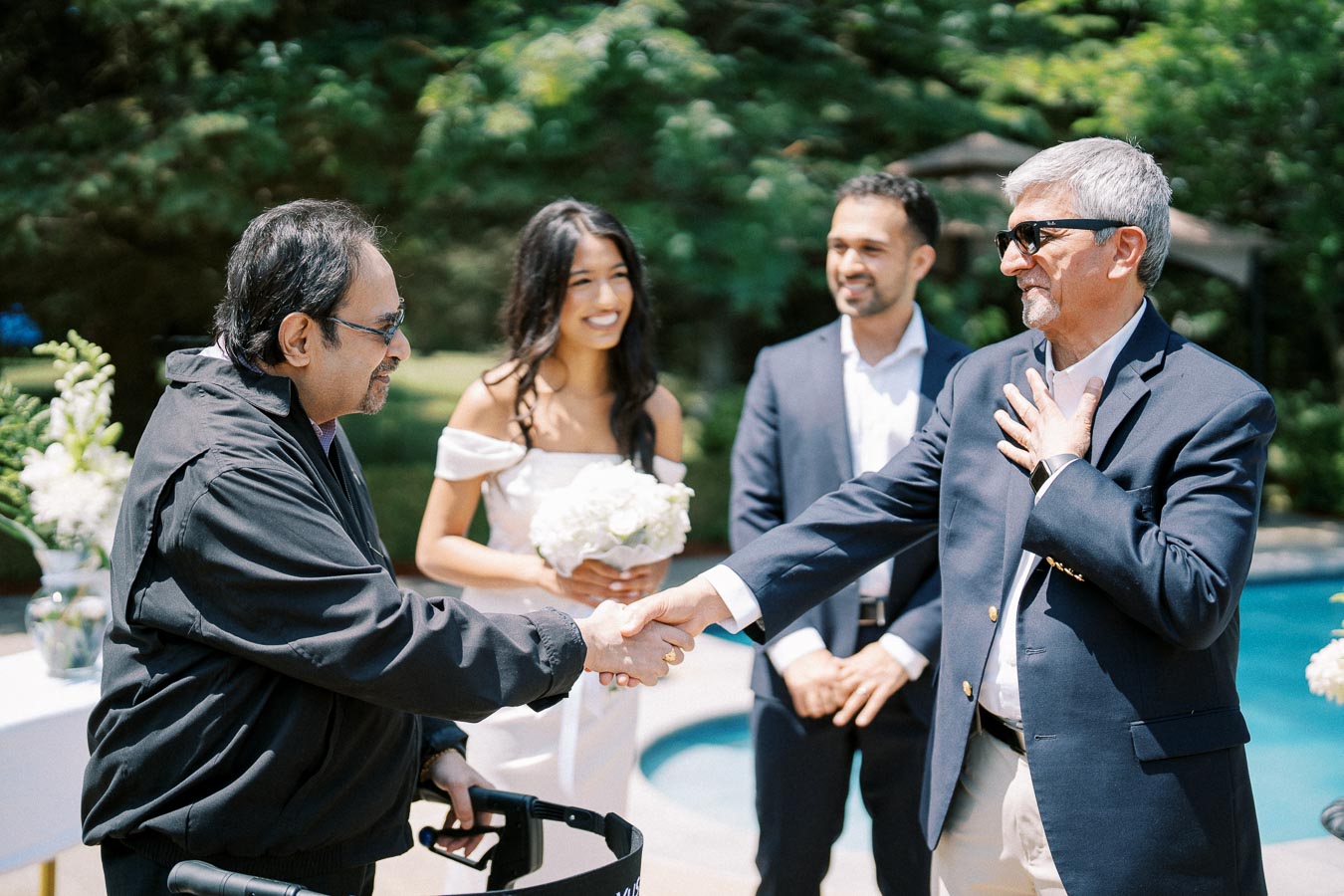 Outdoor wedding ceremony with two men in suits shaking hands, a woman in a white dress holding flowers, and another man in the background, surrounded by greenery and a pool.