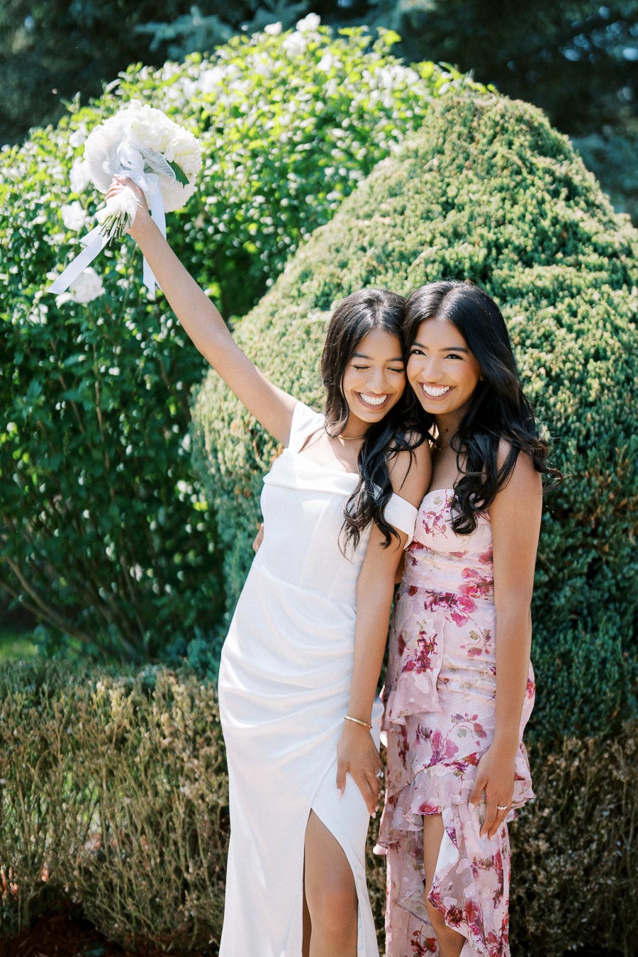 Two women in elegant dresses smiling and posing in a garden, one holding a bouquet.