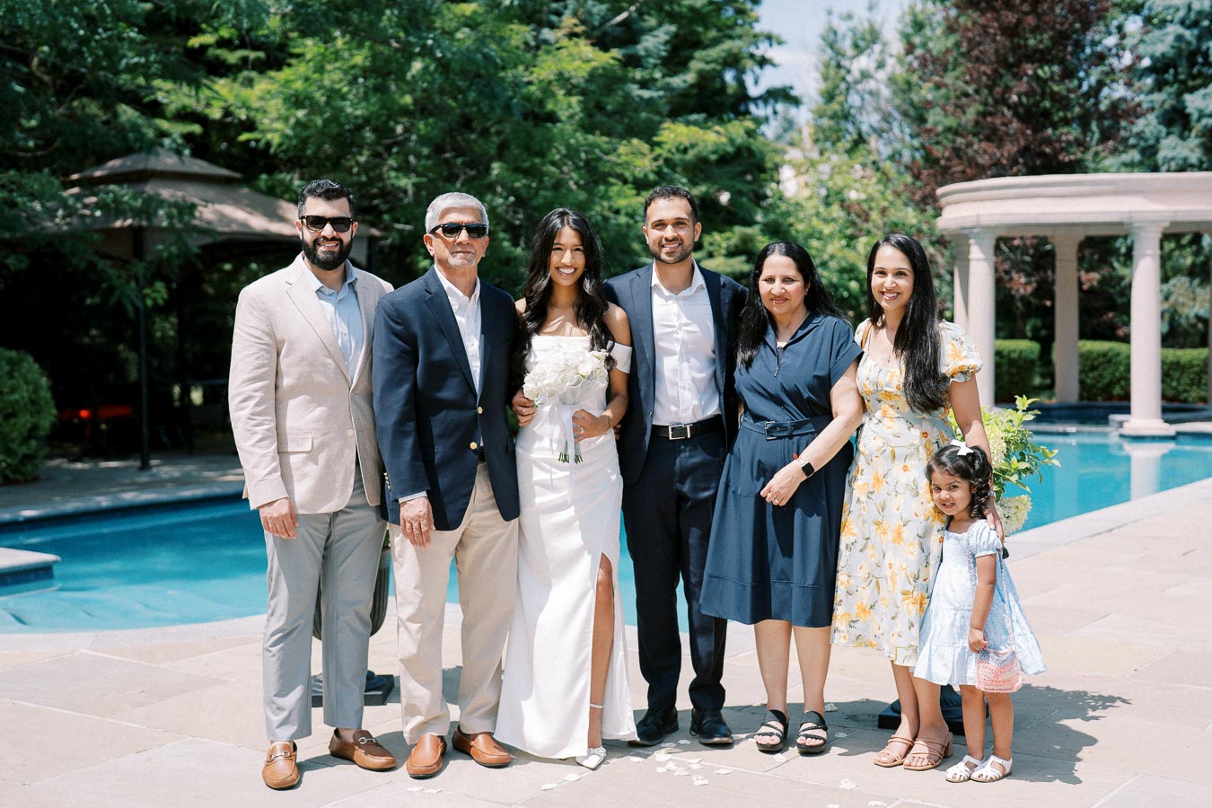 Family group photo at an outdoor wedding, with the bride holding a bouquet, standing by a pool on a sunny day, surrounded by lush greenery and elegant architecture.