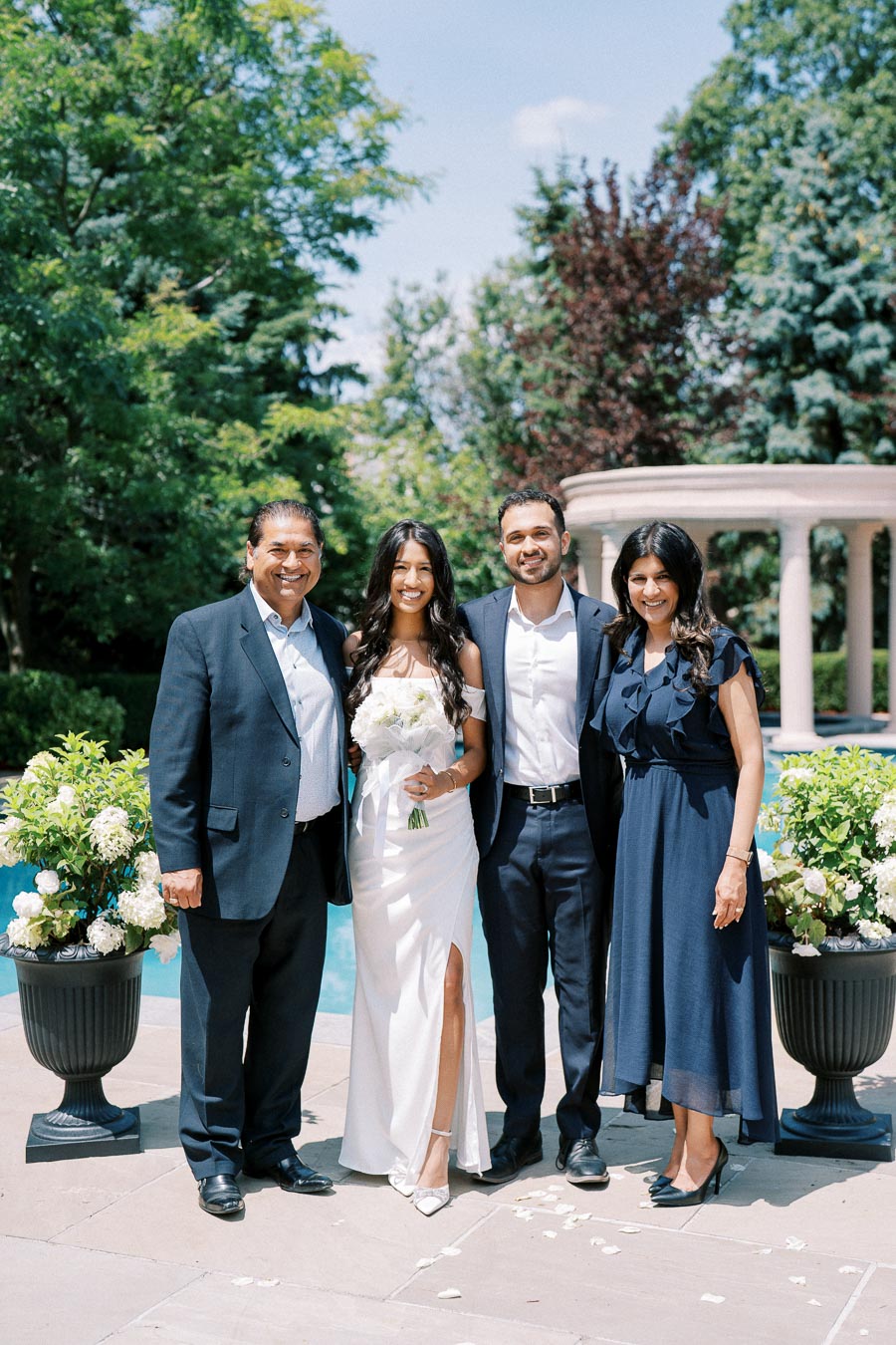 A happy family of four standing together outdoors at a wedding ceremony, with a lush green garden and elegant stone columns in the background. The bride is wearing a white dress and holding a bouquet, surrounded by well-dressed parents and a groom in a suit.