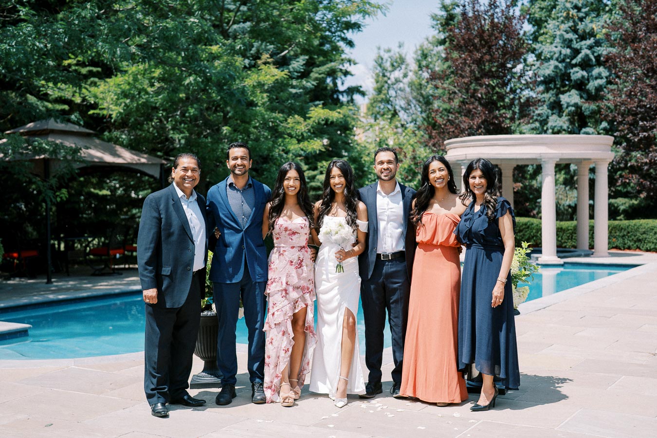 A group of well-dressed people posing together outdoors next to a pool, with lush greenery and a classical white gazebo in the background. The central figure is wearing a white dress, holding a bouquet of flowers, suggesting a celebration or wedding event.