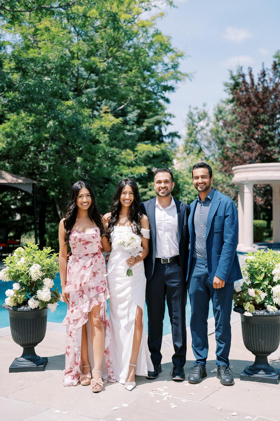 A group of four people posing outdoors at a wedding, with two women in dresses and two men in suits, surrounded by lush greenery and floral arrangements, on a sunny day.