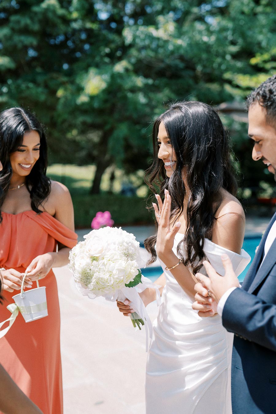 Bride smiling during an outdoor wedding ceremony, holding a bouquet of white flowers, surrounded by guests in formal attire.