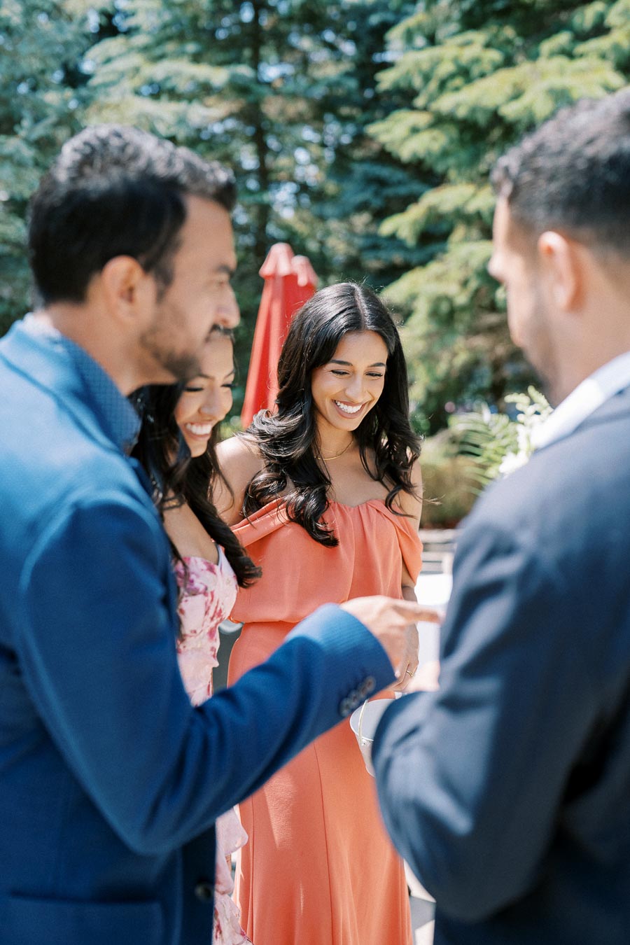 Group of friends enjoying a sunny outdoor gathering, with a woman in a coral dress smiling warmly, surrounded by trees and greenery.