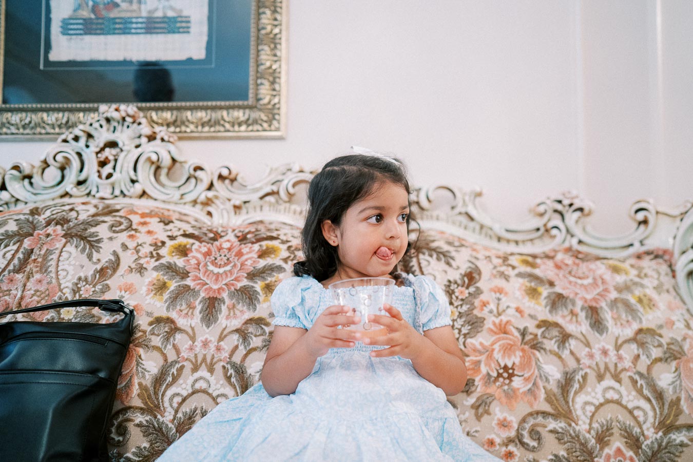 Little girl in a blue dress sitting on an ornate floral sofa, holding a plastic cup with a playful expression.