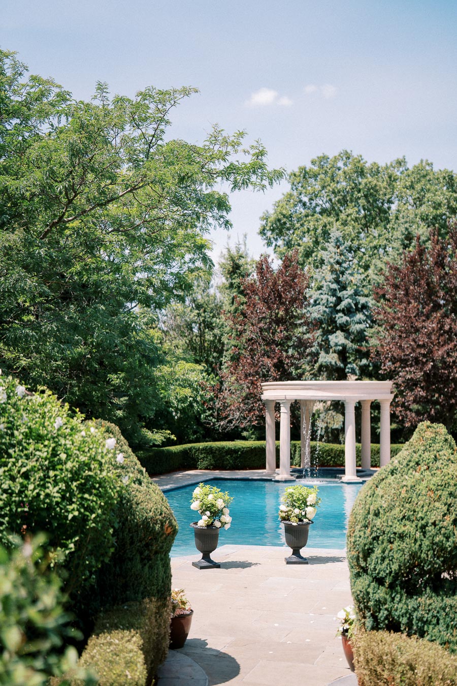 Elegant garden with a stone pathway leading to a serene swimming pool surrounded by lush greenery and white decorative columns. Beautifully manicured shrubs and potted plants enhance the tranquil outdoor setting.