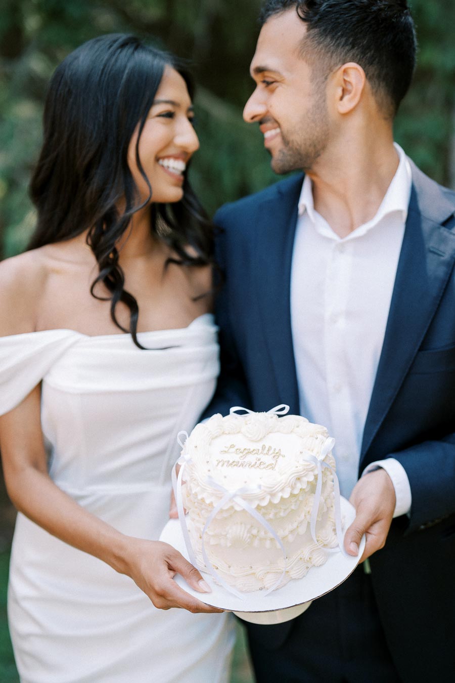 A joyful couple in wedding attire holding a decorated cake with the words Legally Married outside, signifying celebration and union.