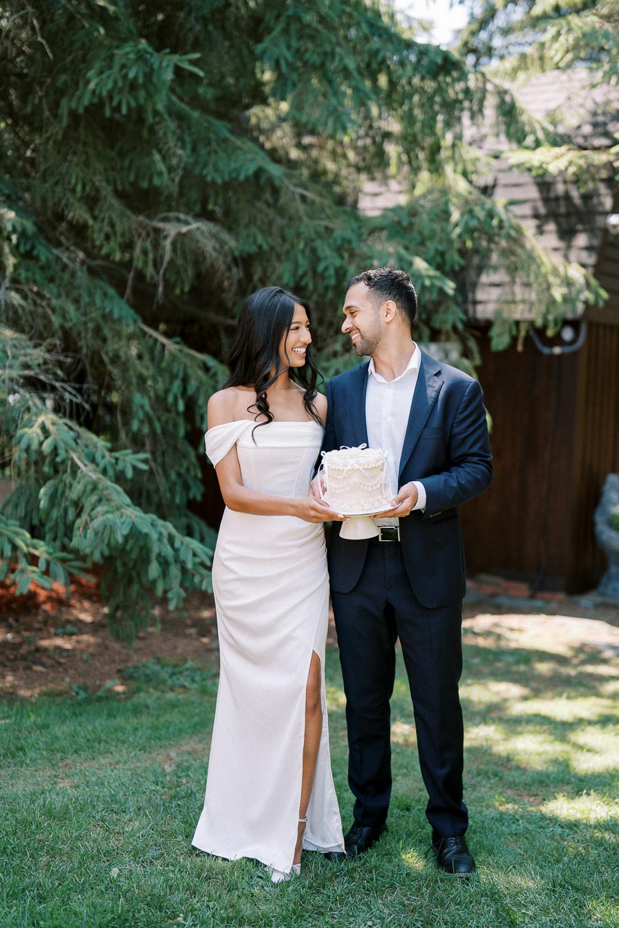 A couple celebrating outdoors in formal attire, holding a white cake with decorative frosting, surrounded by lush greenery and trees.