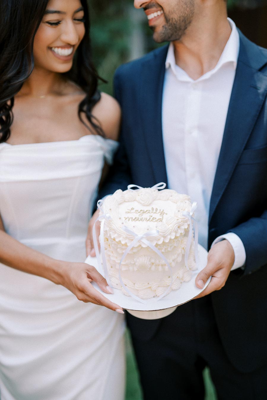 A newlywed couple in wedding attire holds a small, elegantly decorated white cake with the text Legally Married, capturing a joyful wedding celebration moment.