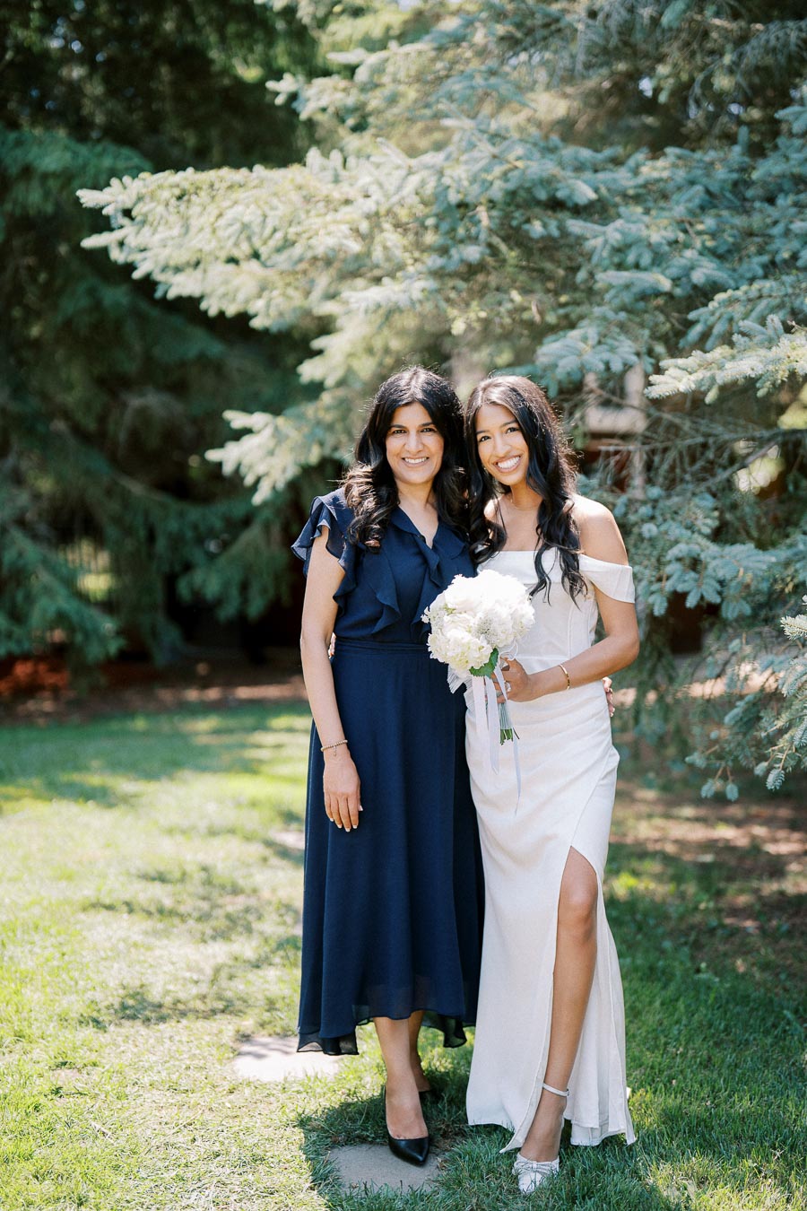 Two women smiling outdoors; one in a blue dress, the other in a white dress holding a bouquet, standing under a pine tree on lush green grass.