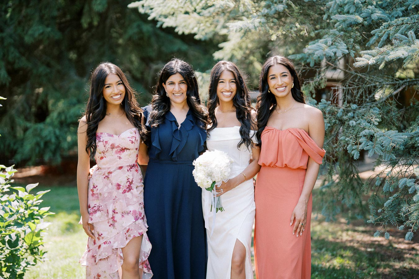 Four women in colorful dresses pose outdoors, surrounded by lush greenery, celebrating a joyful occasion.