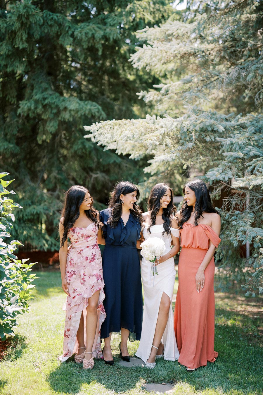 A group of four women in elegant dresses smiling and posing together outdoors, surrounded by lush green trees, creating a warm and joyful wedding atmosphere.