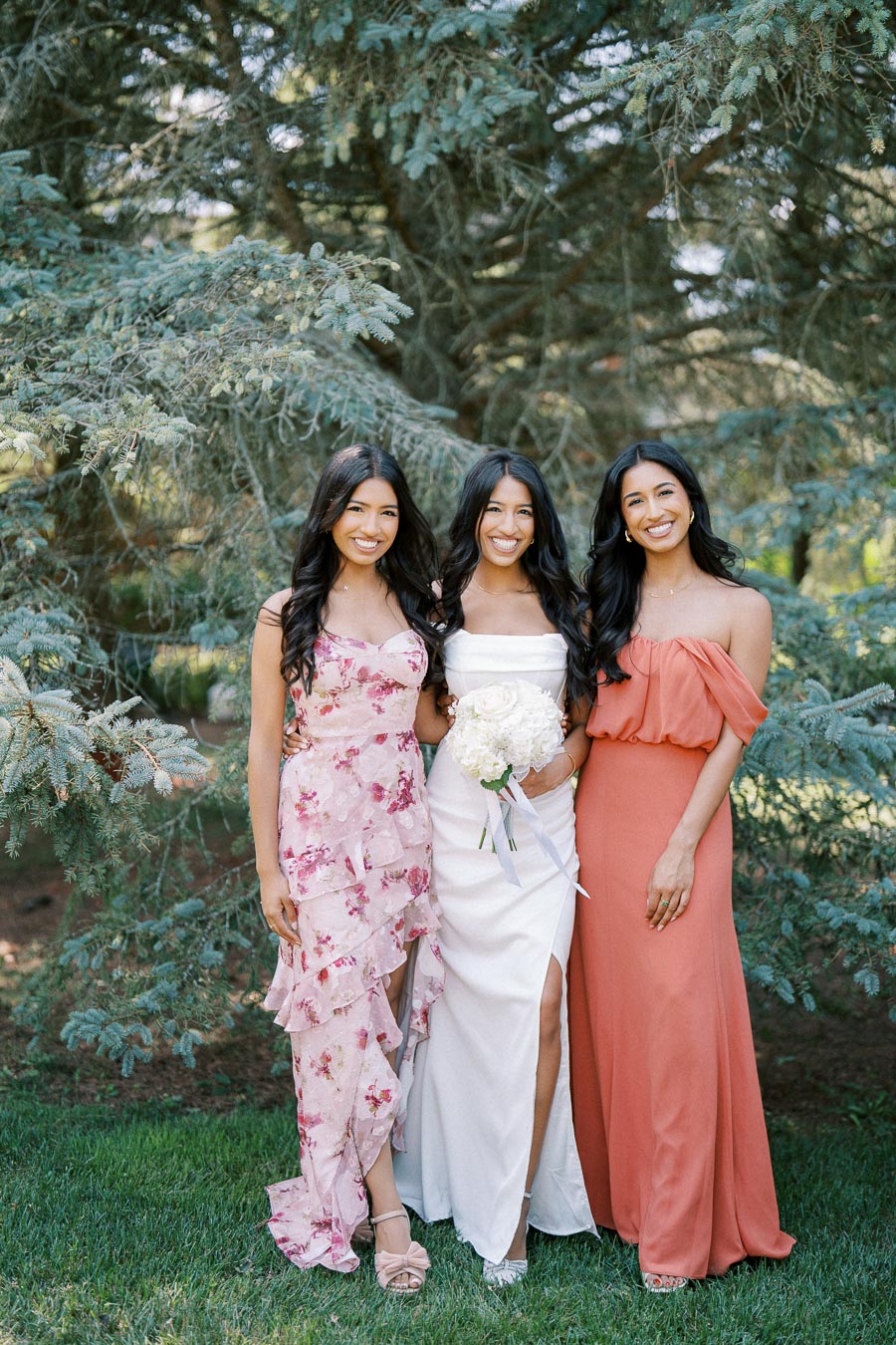 Three women in elegant dresses posing outdoors in front of lush greenery; one in a white gown holding a bouquet, flanked by companions in floral and coral dresses, all smiling.