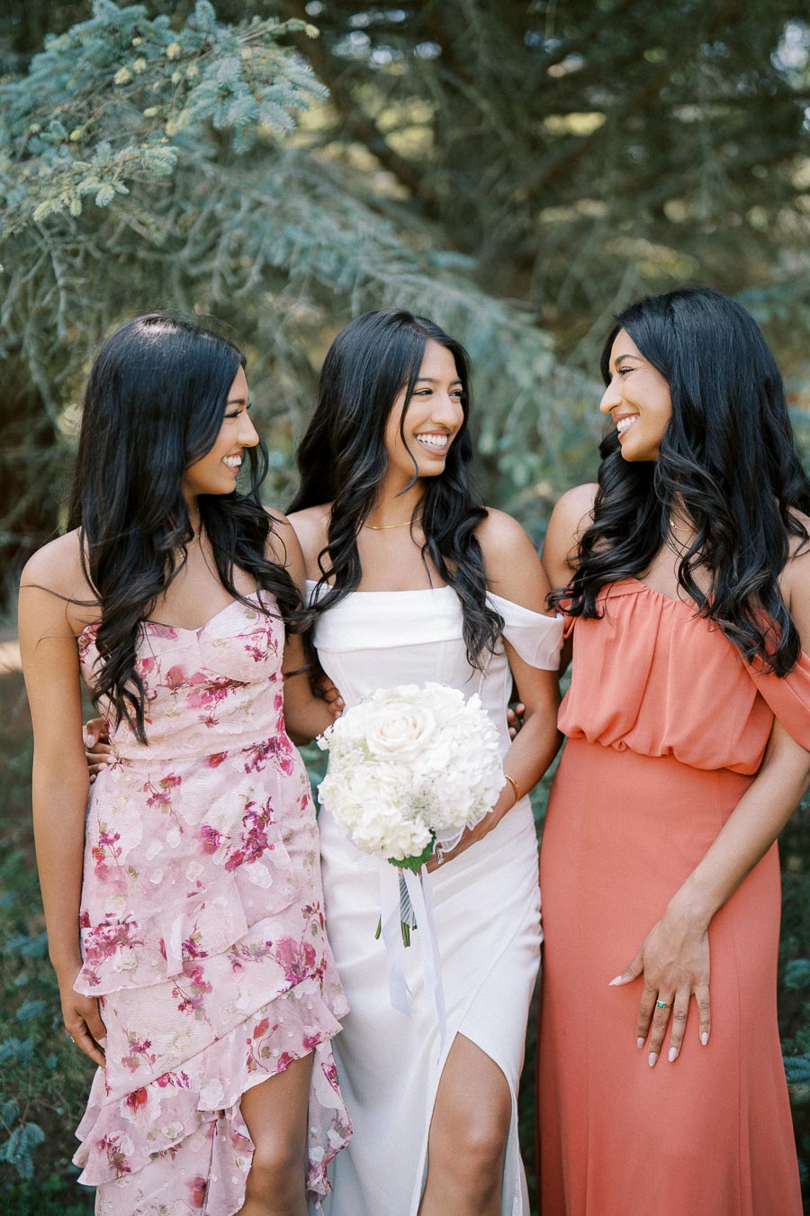 Three women smiling in elegant dresses, standing outdoors surrounded by greenery. One holds a bouquet of white flowers, suggesting a bridal or special occasion photo.
