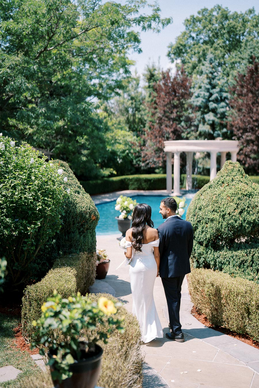 Couple walking through lush garden towards elegant poolside setting with vibrant greenery and classical architecture.