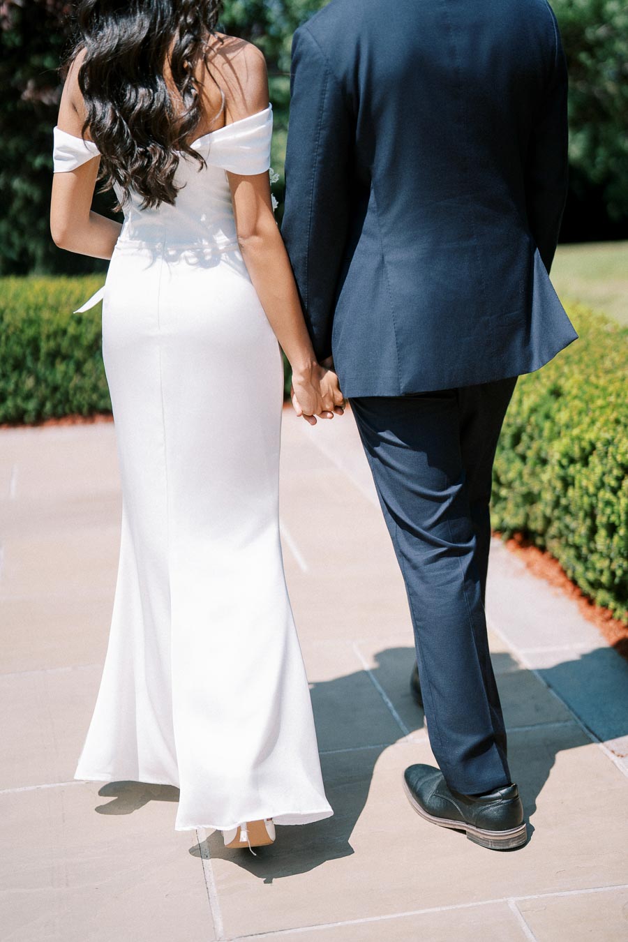Wedding couple walking hand in hand down a sunlit garden path, with the bride in an elegant white gown and the groom in a formal dark suit, surrounded by lush greenery.