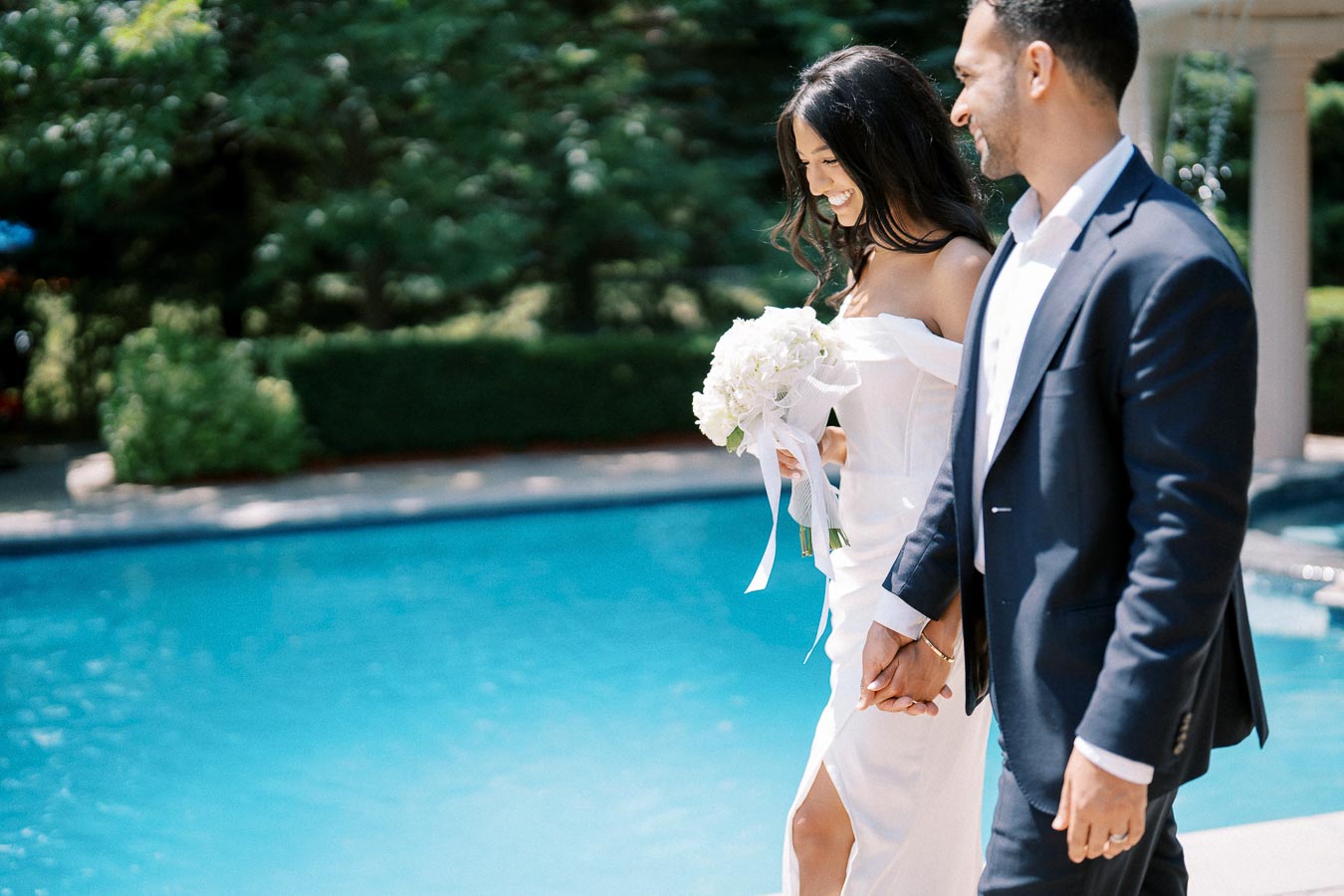 A bride in a white dress and a groom in a suit happily holding hands while walking beside a blue pool, surrounded by lush greenery.