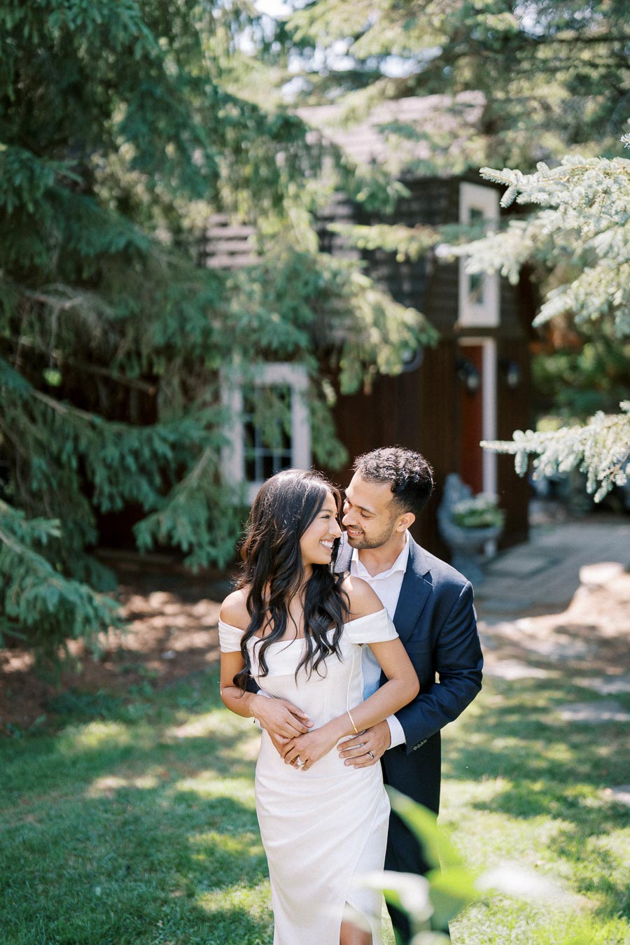 A couple embracing in an outdoor garden setting, surrounded by lush greenery and trees, with a rustic wooden building in the background. The woman is wearing a white dress, and the man is in a navy suit. They are smiling at each other, conveying a sense of joy and romance.